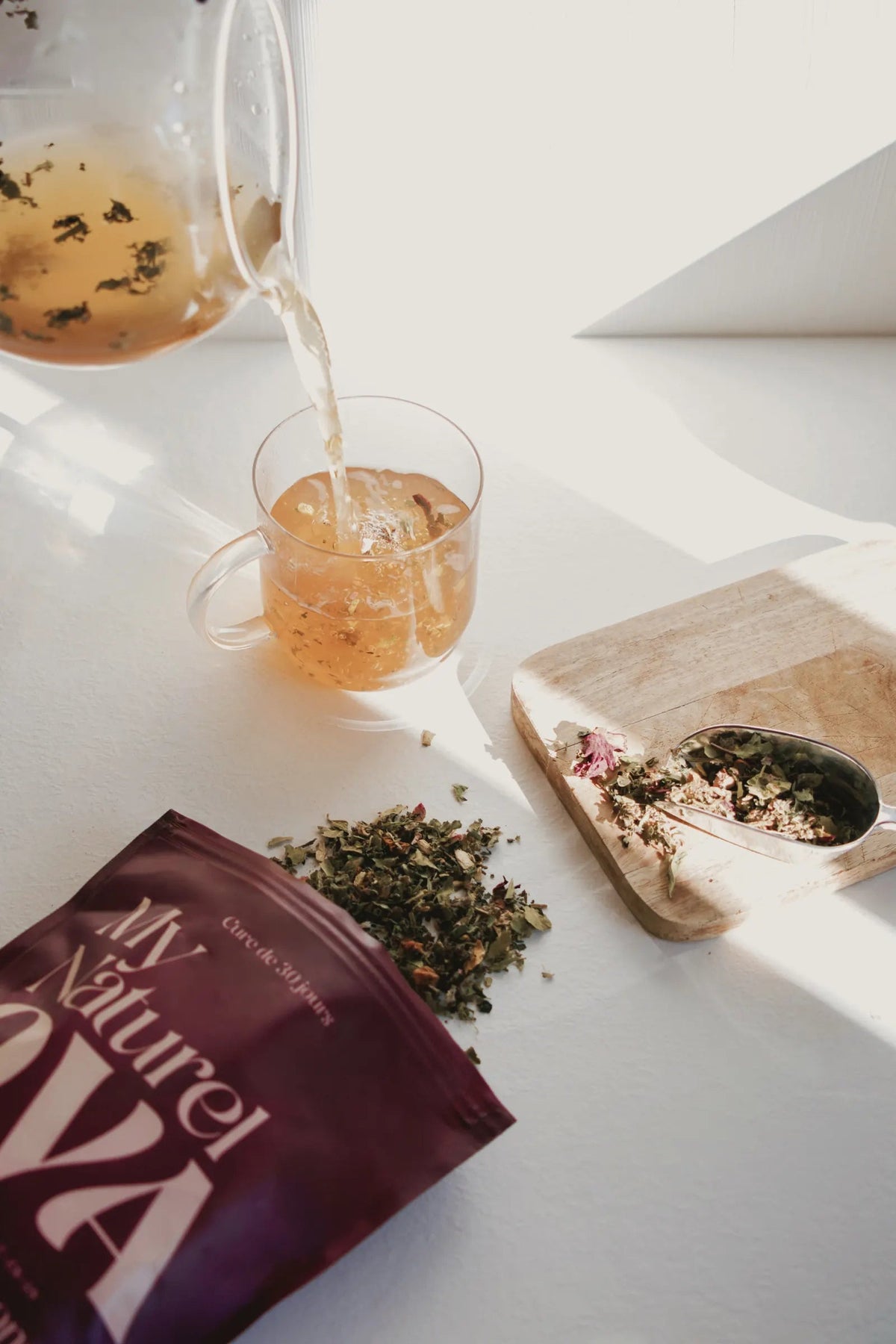 Une infusion dorée versée dans une tasse en verre, le sachet et des plantes au premier plan