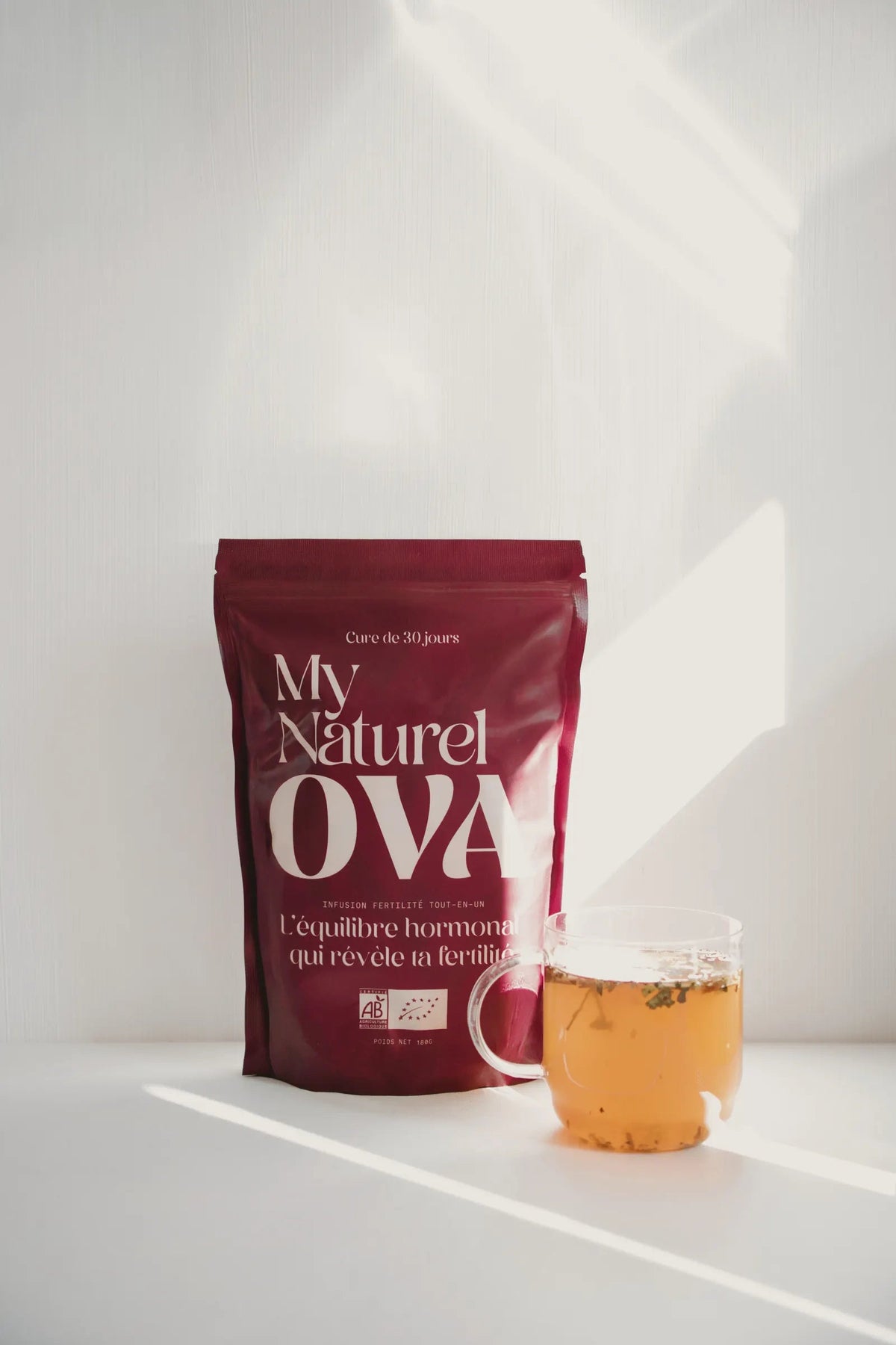 Le sachet My Naturel Ova et une tasse d'infusion dorée dans la lumière